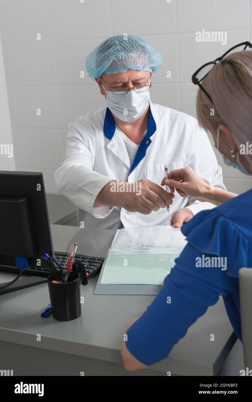 A doctor in a white coat, mask and cap in a medical office sitting at a table gives a pen to a patient to sign an informed consent Stock Photo