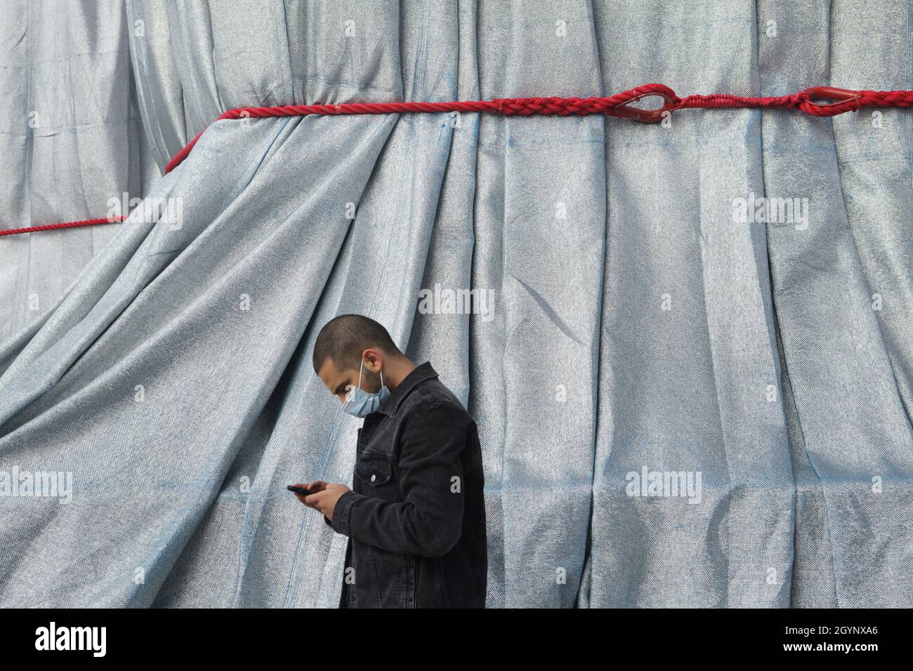 Young man wearing a face mask uses a smartphone in front of the Arc de Triomphe wrapped in silver-blue fabric fastened with red ropes in the Place Charles de Gaulle in Paris, France. The Arc de Triomphe was wrapped for two weeks being converted to an artwork as it was designed by Christo and Jeanne-Claude in September 2021. Stock Photo