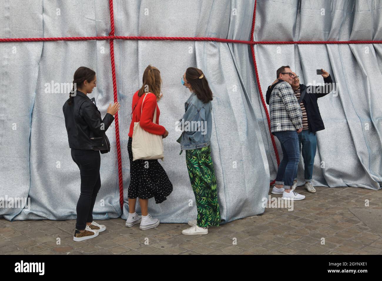 Couple uses a smartphone to make a selfie in front of the Arc de Triomphe wrapped in silver-blue fabric fastened with red ropes in the Place Charles de Gaulle in Paris, France. The Arc de Triomphe was wrapped for two weeks being converted to an artwork as it was designed by Christo and Jeanne-Claude in September 2021. Stock Photo