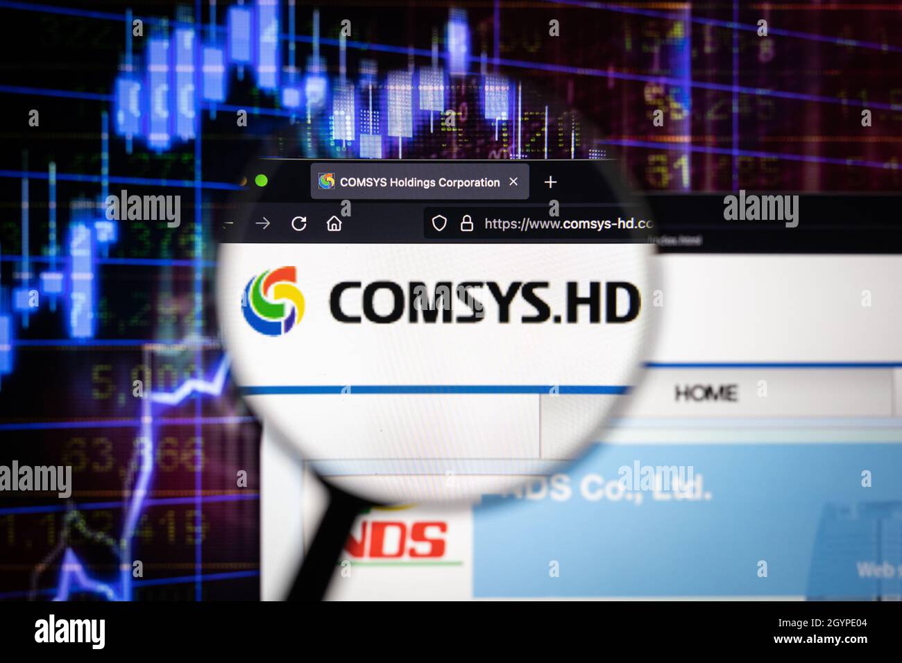 Comsys.HD company logo on a website with blurry stock market graphs in the background, seen on a computer screen through a magnifying glass. Stock Photo