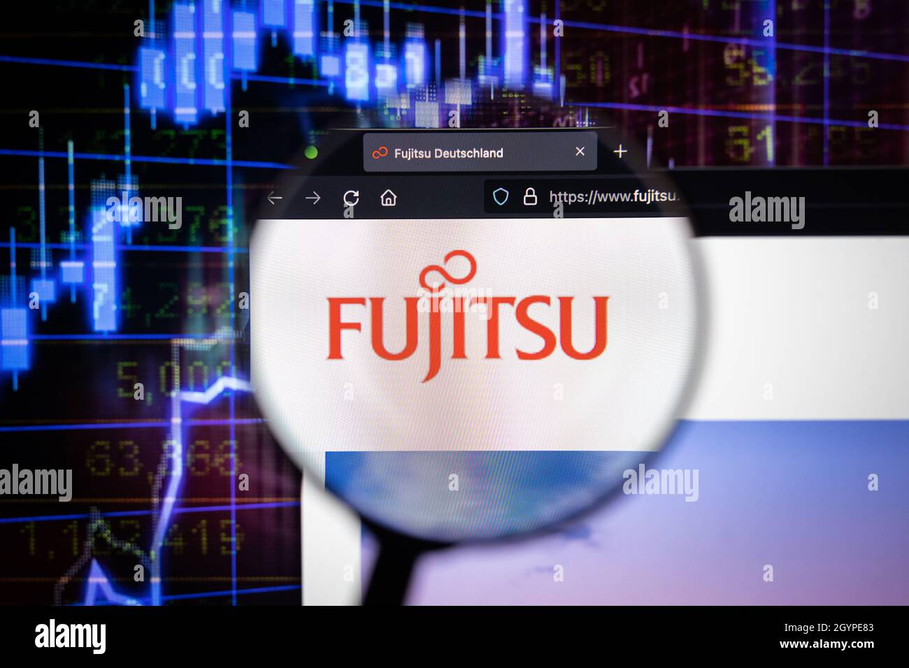 Fujitsu company logo on a website with blurry stock market graphs in the background, seen on a computer screen through a magnifying glass. Stock Photo