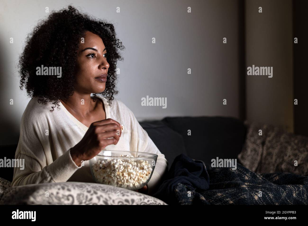 horizontal portrait of an african american woman sitting on a sofa. Face is lit up with TV light. She is holding a bowl of popcorn Stock Photo