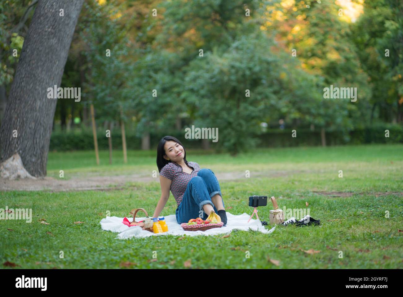 Autumn picnic at city park - lifestyle portrait of young happy and beautiful Asian Chinese woman sitting grass with blanket and fruit basket enjoying Stock Photo