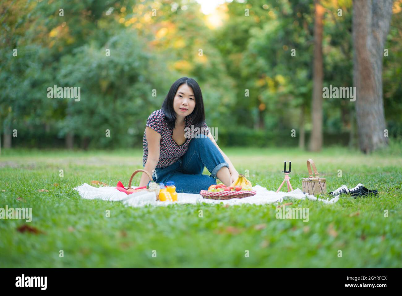 Autumn picnic at city park - lifestyle portrait of young happy and beautiful Asian Korean woman sitting grass with blanket and fruit basket enjoying r Stock Photo