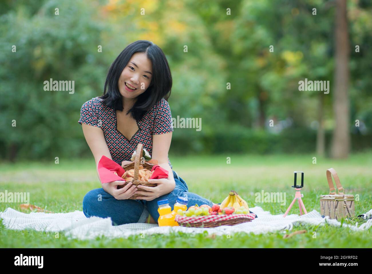 Autumn picnic at city park - lifestyle portrait of young happy and beautiful Asian Korean woman sitting grass with blanket and fruit basket enjoying r Stock Photo