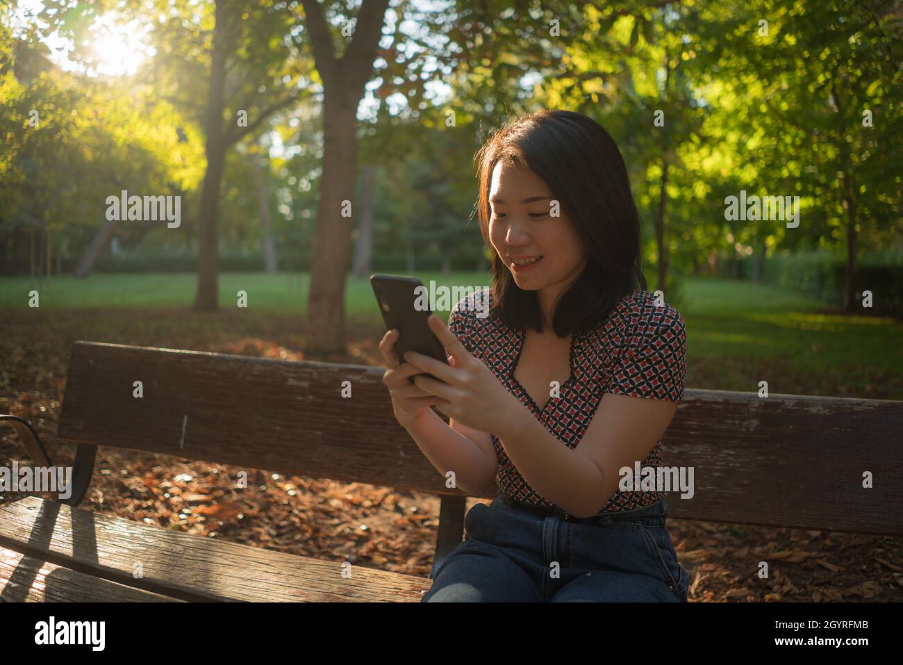 lifestyle portrait of young happy and beautiful Asian Chinese woman using internet app on mobile phone relaxed and cheerful at city park enjoying natu Stock Photo