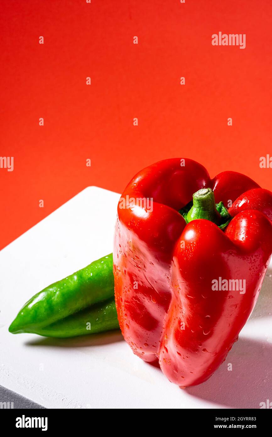 Photograph of a red pepper and a green Italian pepper on a white bench and red background.The photo is shot from a top point of view and taken in vert Stock Photo