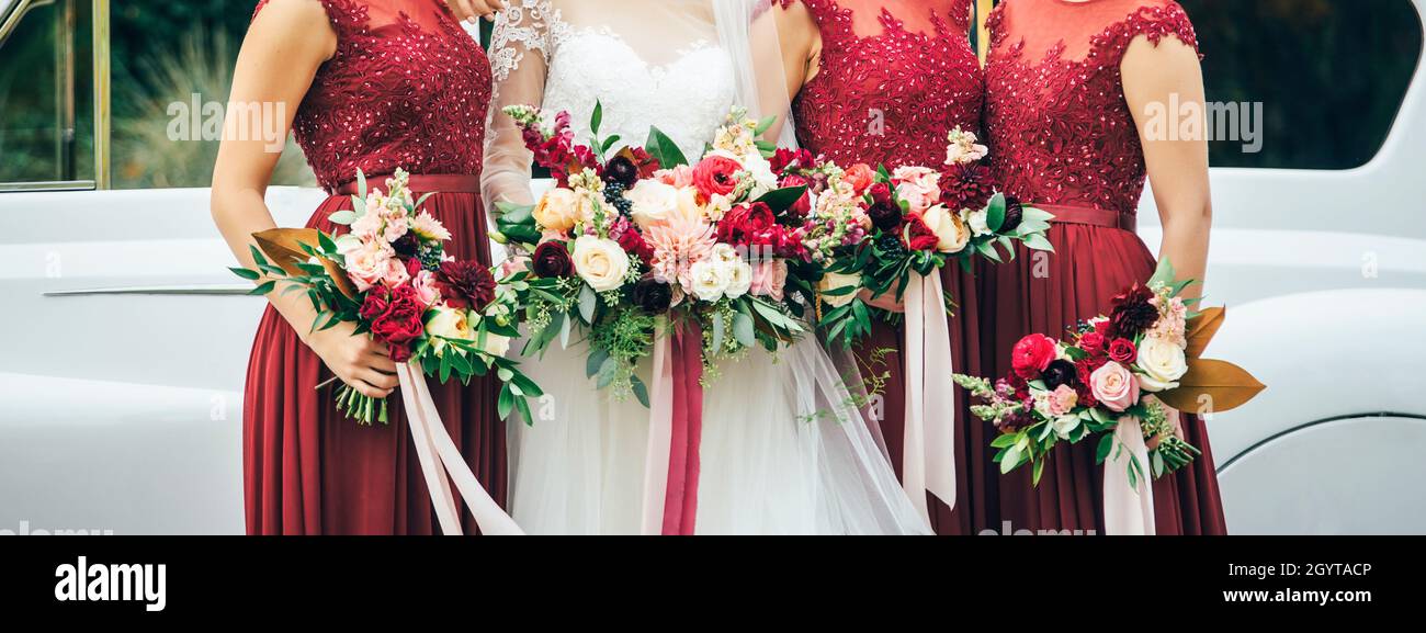 bride in white wedding dress and bridesmaids in red dresses are staying near the old white car with beige pink yellow and red flowers Stock Photo