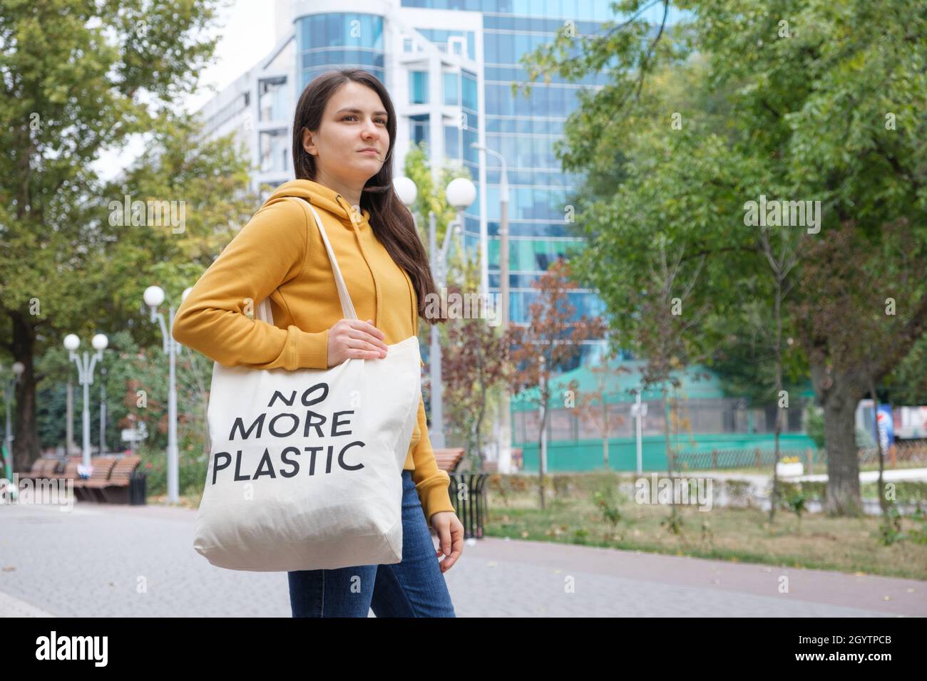 A brunette woman in an orange hoodie walks with a fabric bag that says No more plastic in the city Stock Photo