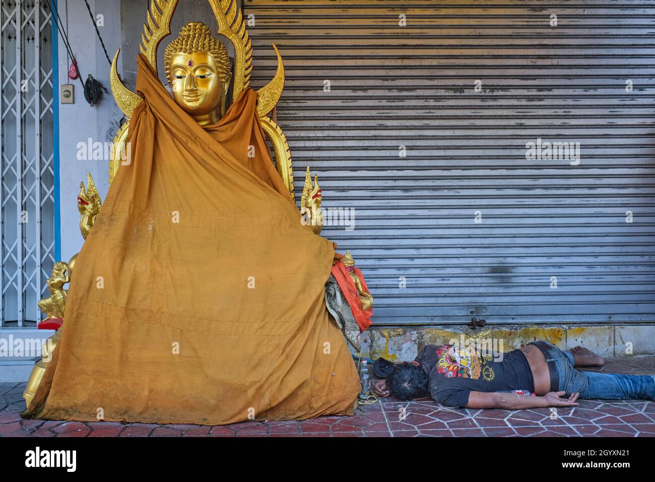 An apparently drunk man sleeps next to a Buddha statue placed outside a shop for Buddhist paraphernalia; Bamrung Muang Road, Bangkok, Thailand Stock Photo