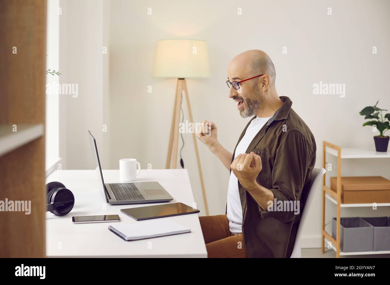 Joyful and happy adult man emotionally rejoices sitting in front of laptop in his home office. Stock Photo