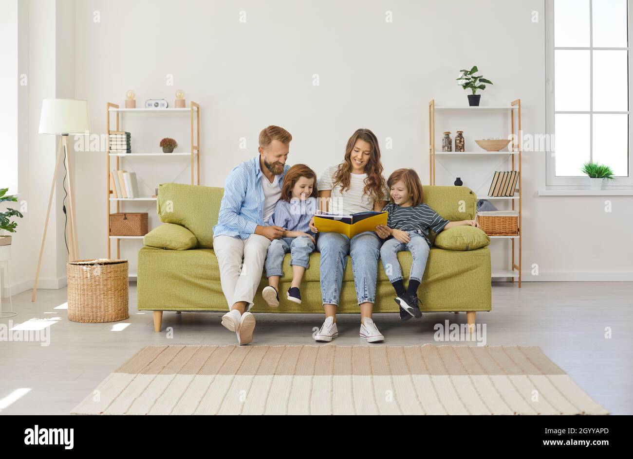 Happy parents and kids reading book while sitting together on sofa in modern living room Stock Photo