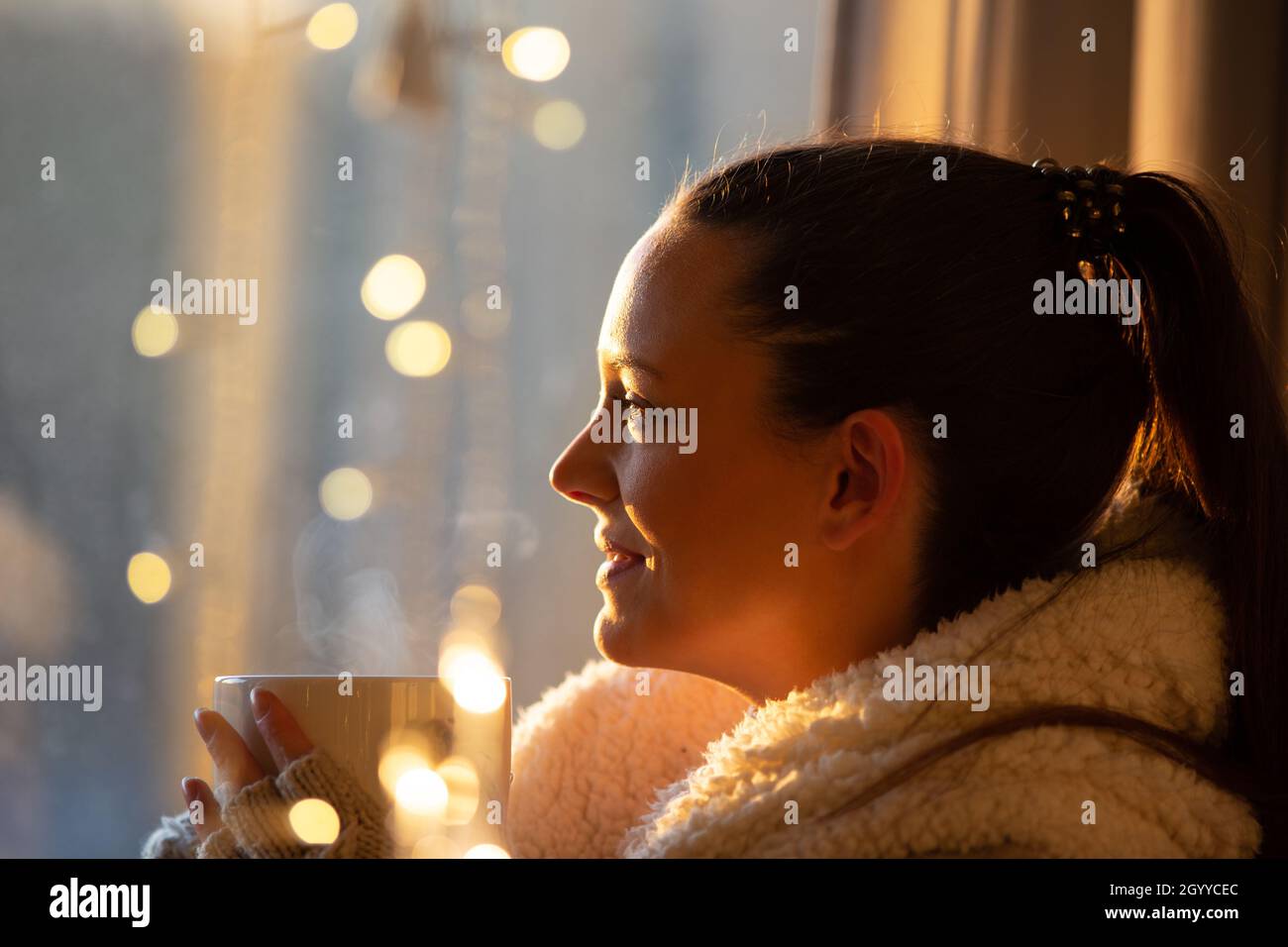 Beautiful girl covered with blanket holding cup of hot coffee with christmas lights in background, Festive moments concept Stock Photo