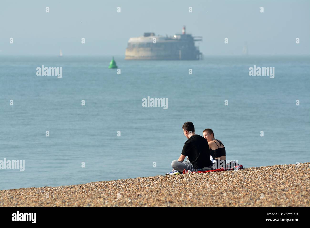 A couple of people sat on the shingle of Southsea beach, Portsmouth, England. The Victorian Spitbank fort in the distance Stock Photo