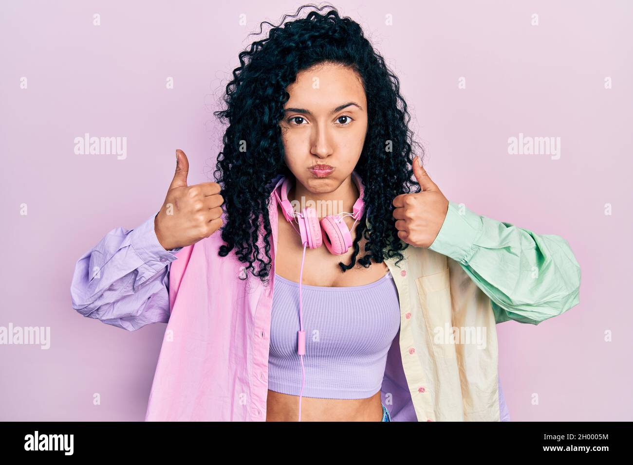 Young hispanic woman with curly hair doing thumbs up positive gesture puffing cheeks with funny face. mouth inflated with air, catching air. Stock Photo