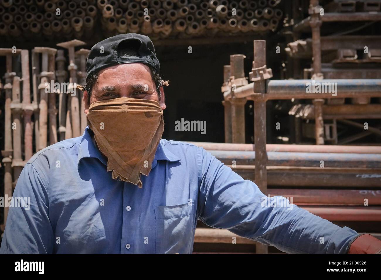 Mature middle eastern migrant laborer wearing wearing blue uniform and dirty mask working for scrap metal supply warehouse for recycling  Stock Photo