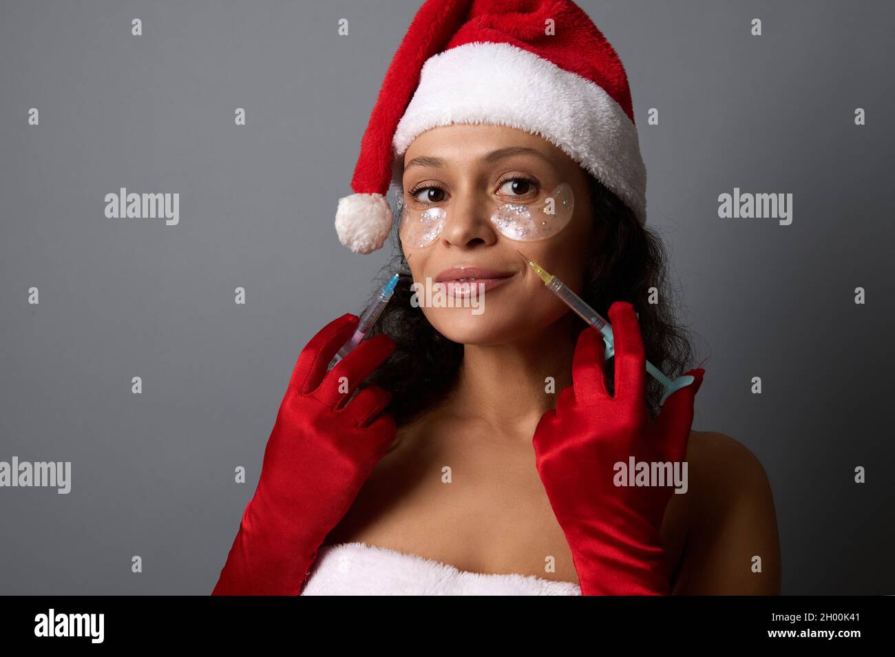 Gorgeous woman dressed in Santa for Christmas holidays, holds syringes near her face, ready for beauty injection on nasolabial folds. Anti-aging, face Stock Photo