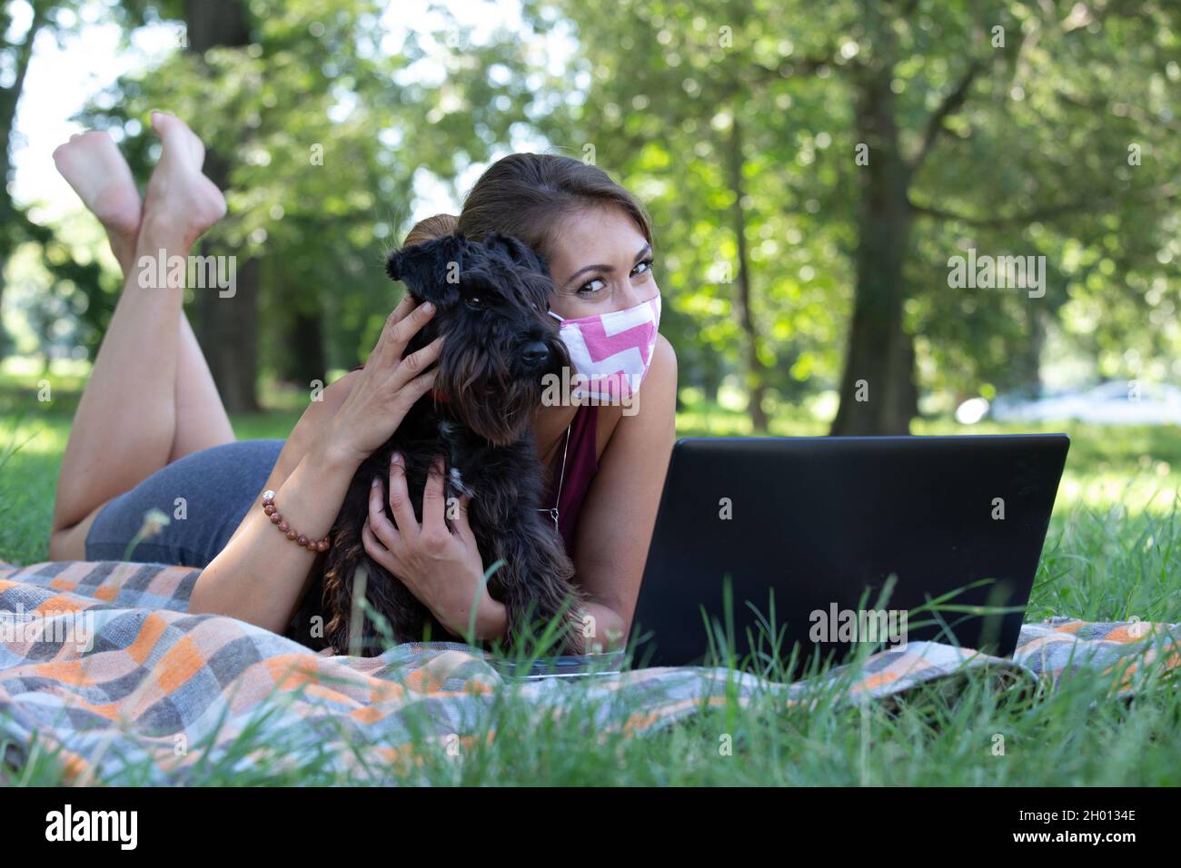 Businesswoman with protective mask working on laptop in park with dog laying next to her Stock Photo