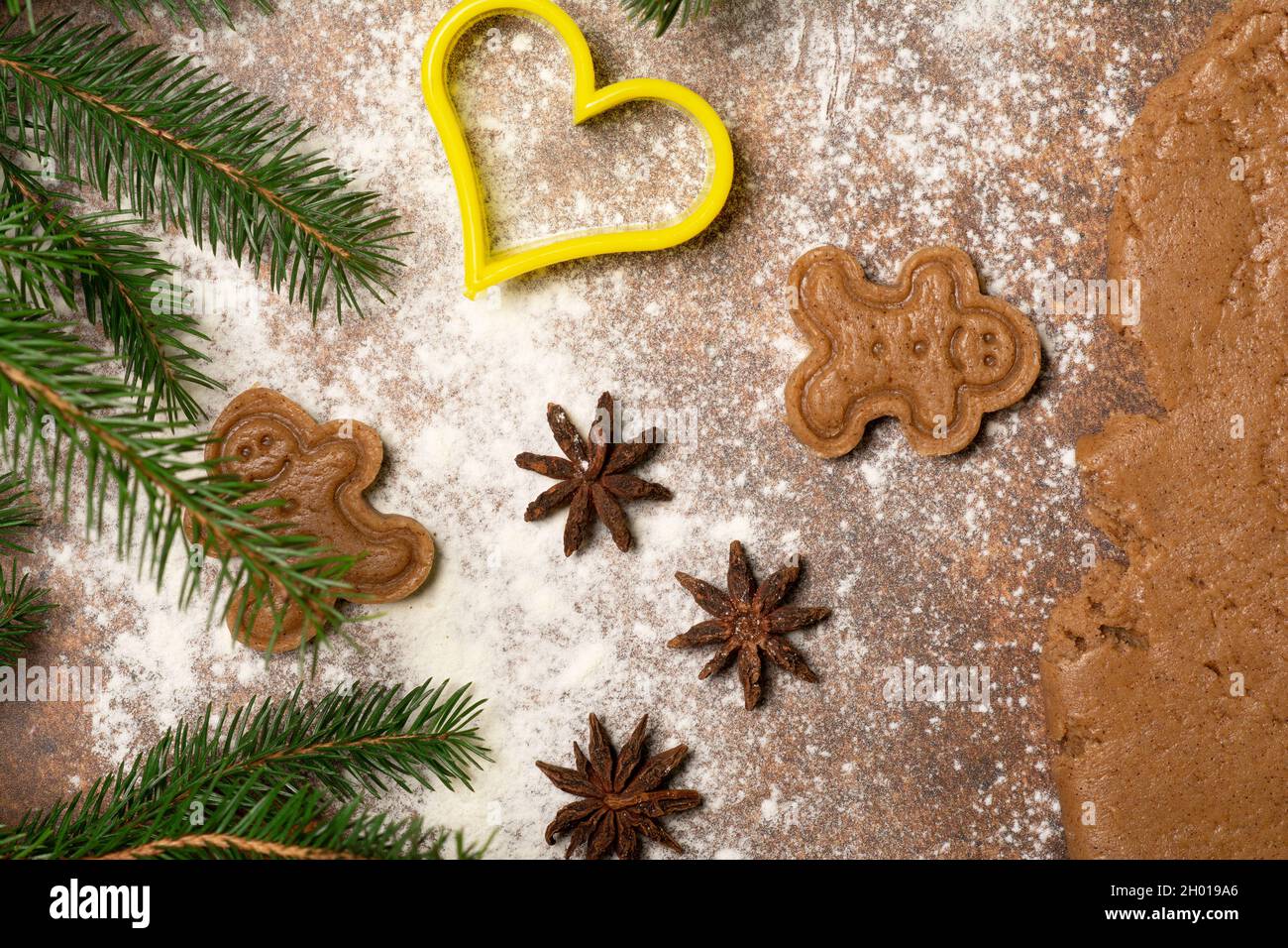 Two gingerbread man cookies, star anise fruits, a cookie cutter, fir branches, a piece of rolled out dough are lying on a surface sprinkled with flour Stock Photo