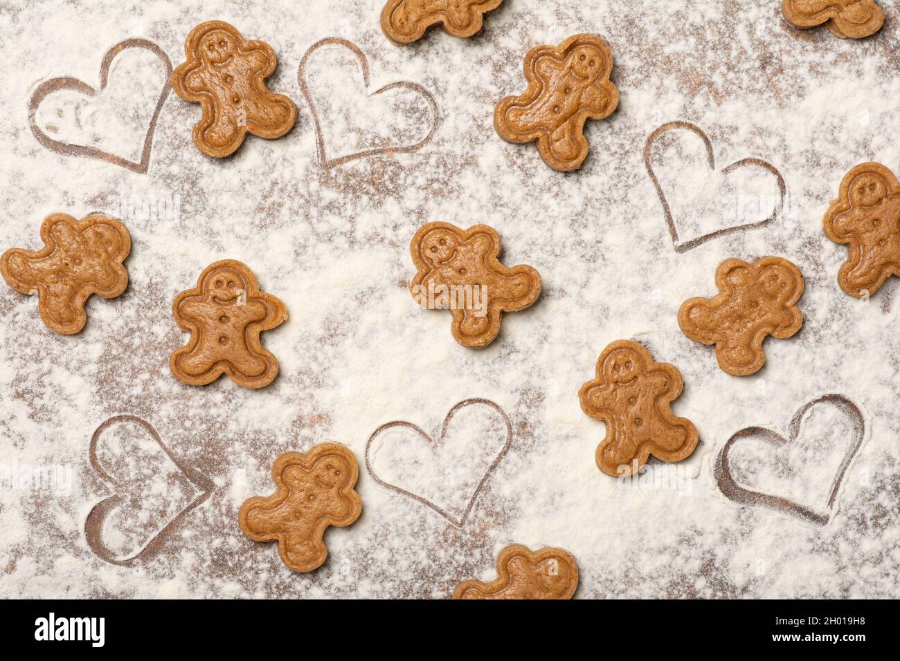 Festive Christmas pattern made of unbaked gingerbread cookies and heart-shaped marks on the kitchen work surface sprinkled with flour. Table top view. Stock Photo