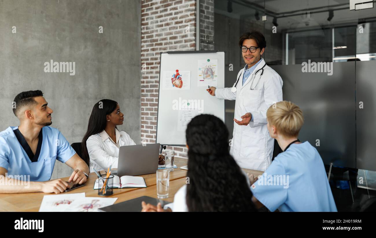 Multiracial team of doctors having morning breefing at clinic, handsome man chief physician making presentation in front of his male and female intern Stock Photo
