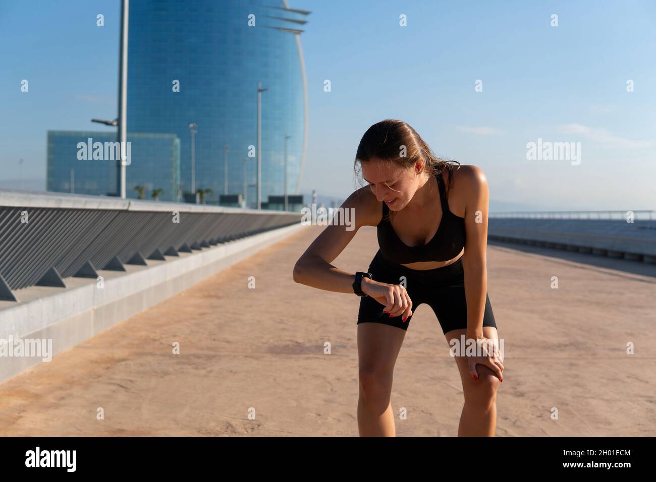 Tired female athlete in sportswear leaning on knee and checking pulse on fitness tracker with smile during break in cardio workout in city. Stock Photo