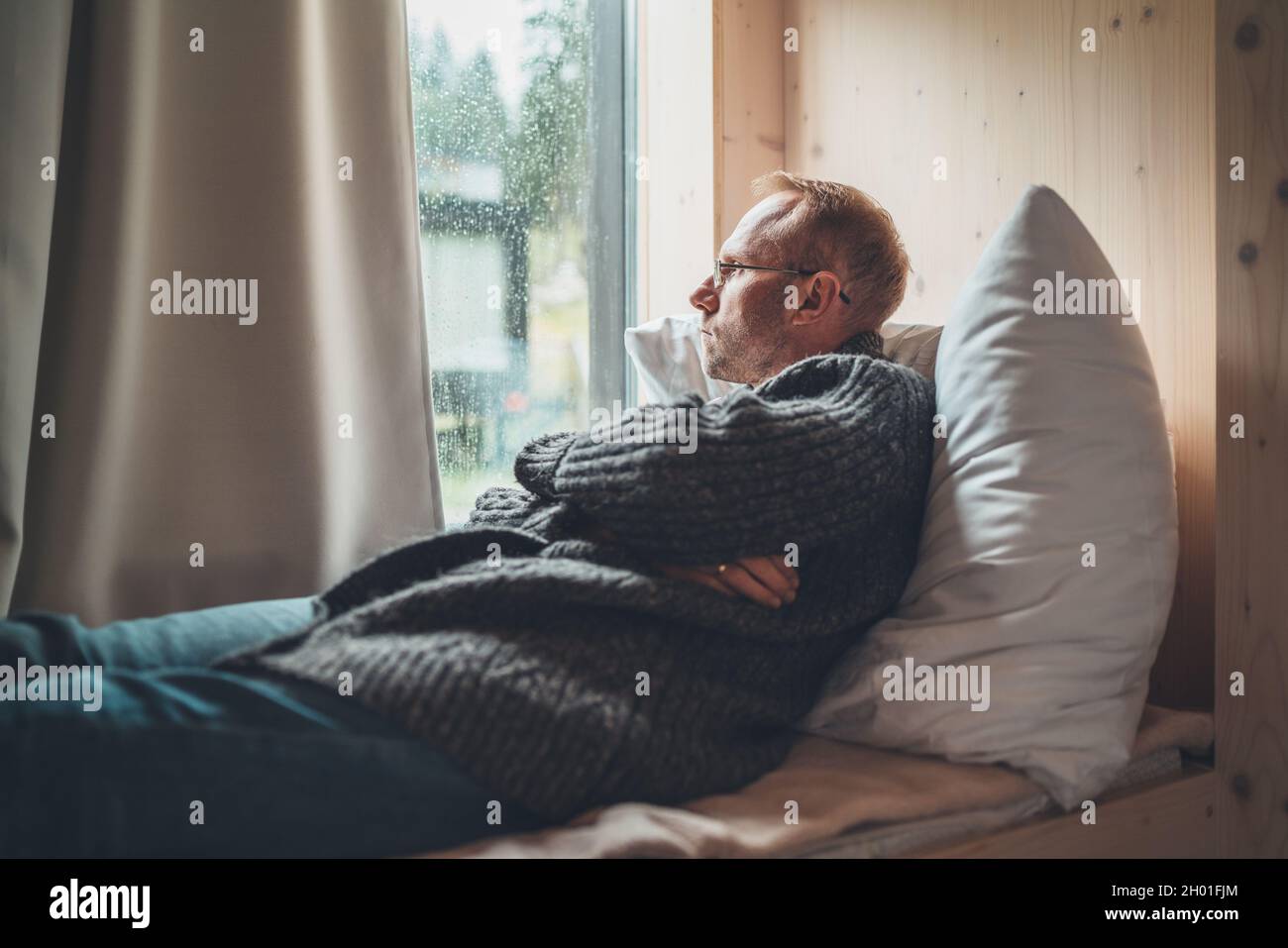 Portrait of thinking sad Middle-aged man in eyeglasses dressed open cardigan lying on cozy bed next to window looking on street through raindrop glass Stock Photo