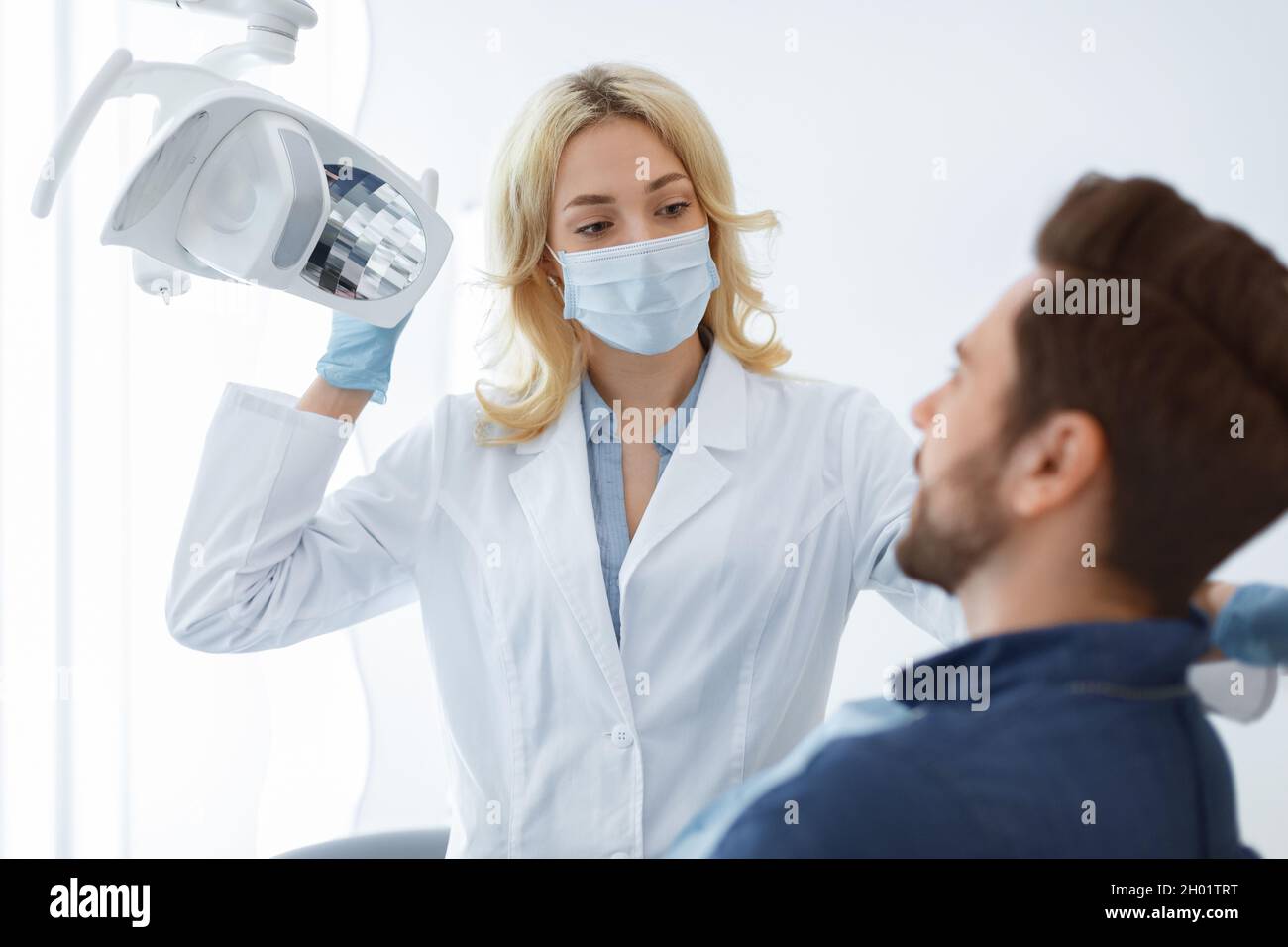 Female dentist blonde woman in face mask and medical gloves greeting male patient sitting in dental chair, turning on lamp while listening to bearded Stock Photo