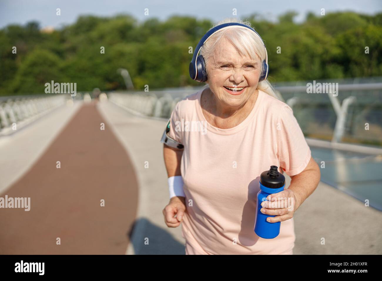 Positive senior lady with headphones runs along bridge in summer Stock Photo