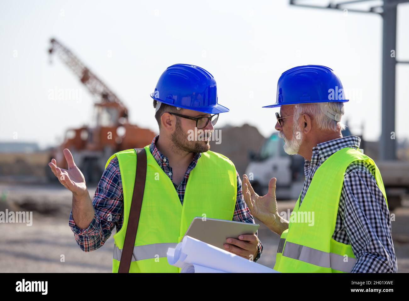 Two engineers with tablet and blueprints talking at building site with metal construction in background Stock Photo