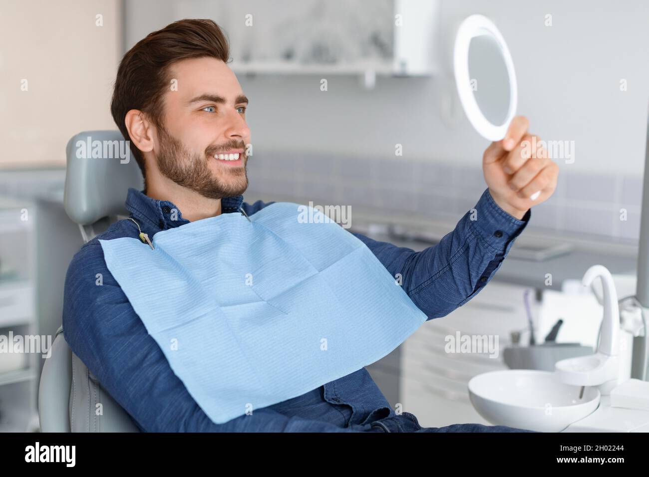 Male patient looking at his beautiful healthy white smile, holding dental hand mirror, happy young man checking results of treatment at modern dentist Stock Photo