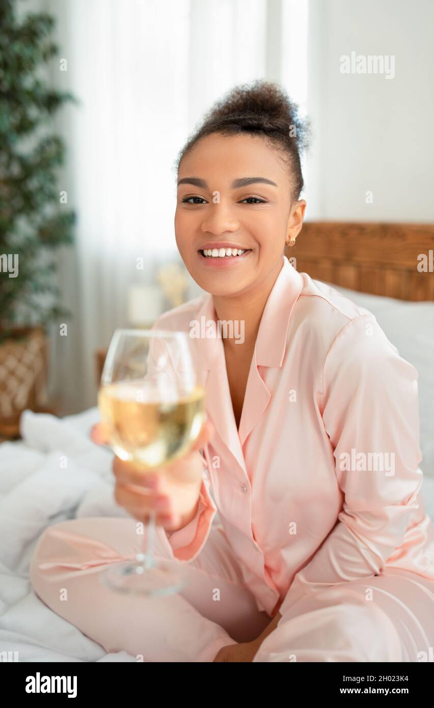 Time for youself concept. Portrait of happy african american lady sitting on bed, holding glass of alcohol beverage, drinking wine and smiling at came Stock Photo