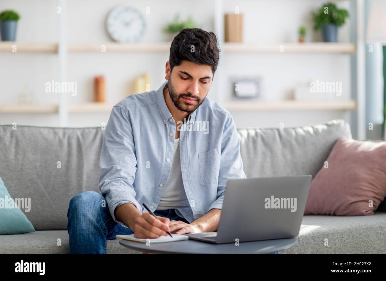 Arab male freelancer writing in notepad and using laptop, taking notes ...