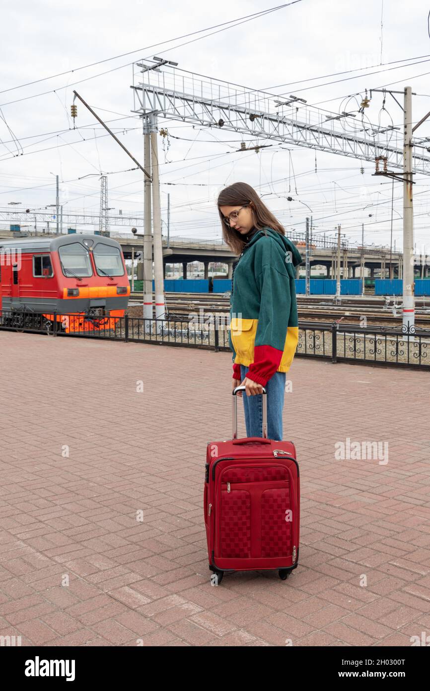 Caucasian girl student in bright clothes with a suitcase going to the electric train at the station. Girl with a suitcase at the train station. A trip Stock Photo