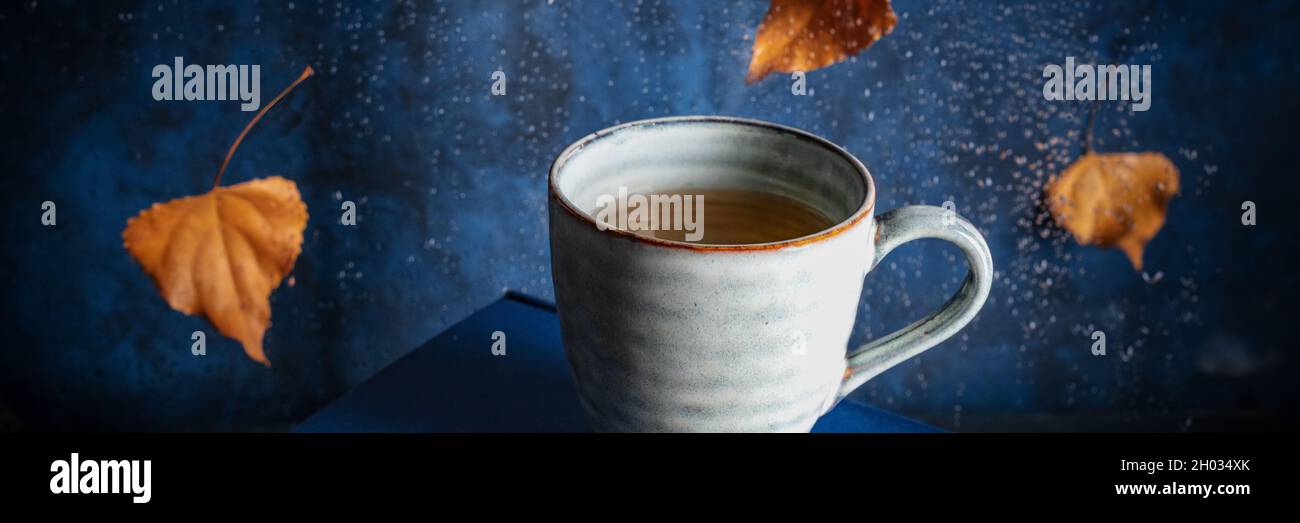 Autumn panorama with a cup of tea and falling leaves behind a window ...