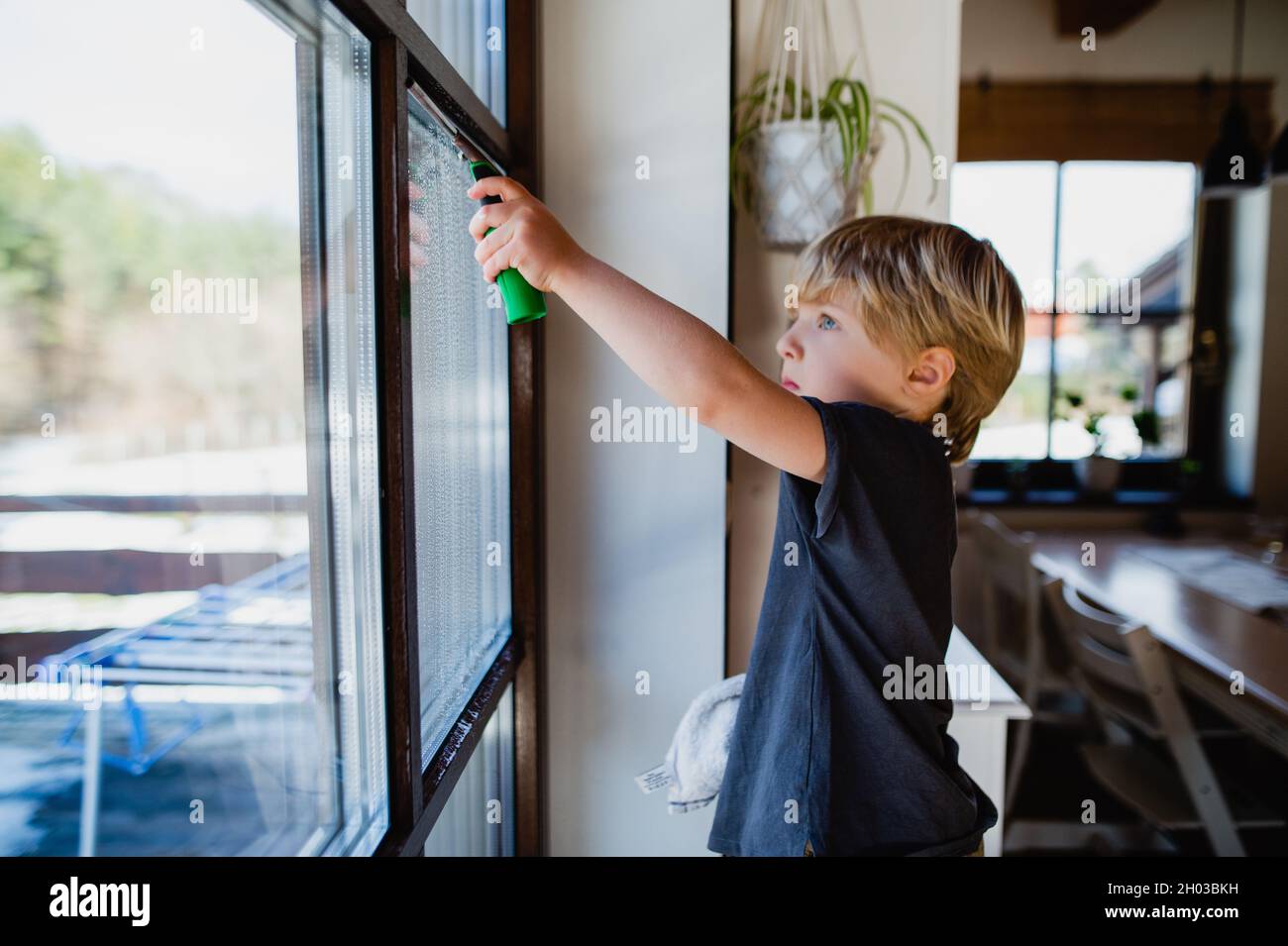 Side view of little boy cleaning windows indoors at home, daily chores