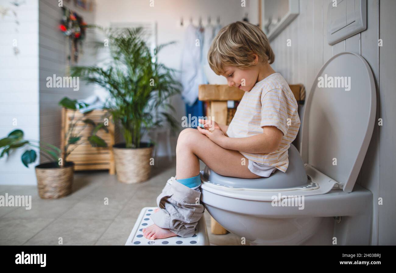Side view of cute small boy sitting on toilet indoors at home, using smartphone. Stock Photo