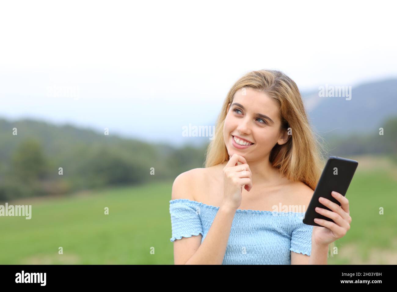 Happy teen holding smart phone and wondering looking at side outdoors in a field Stock Photo