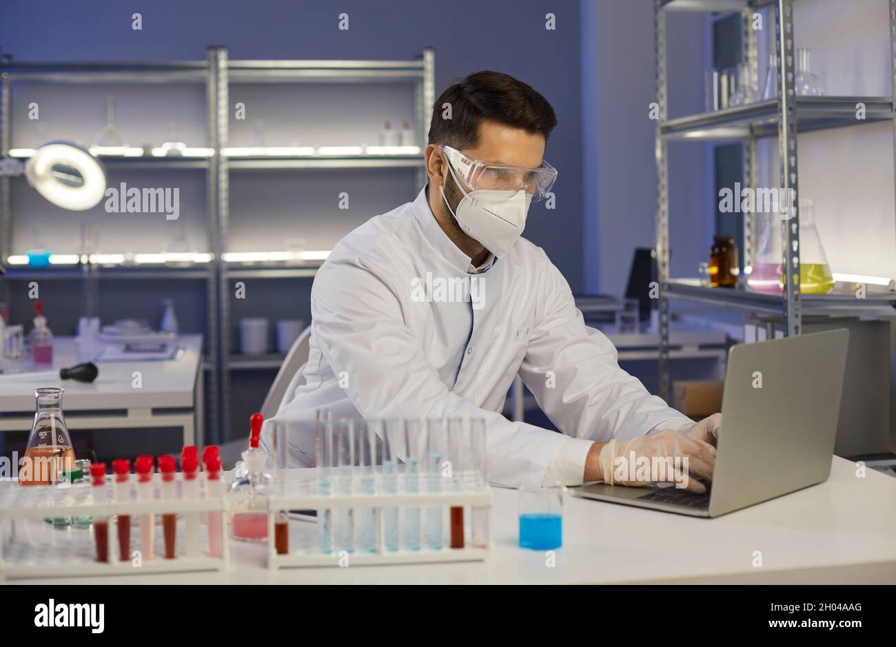 Male scientist researcher in protective respiratory mask typing on laptop Stock Photo