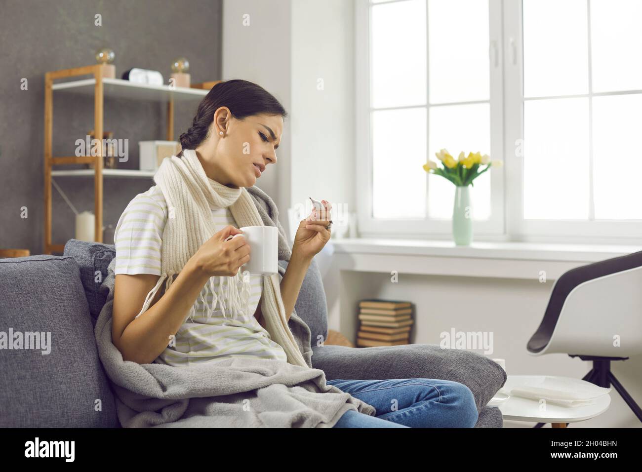 Young woman who has the flu checks her temperature while sitting on a sofa at home. Stock Photo
