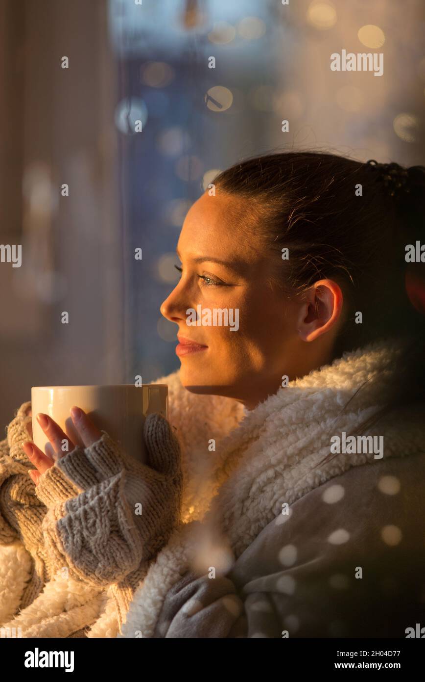 Beautiful girl covered with blanket holding cup of hot coffee with christmas lights in background, Festive moments concept Stock Photo