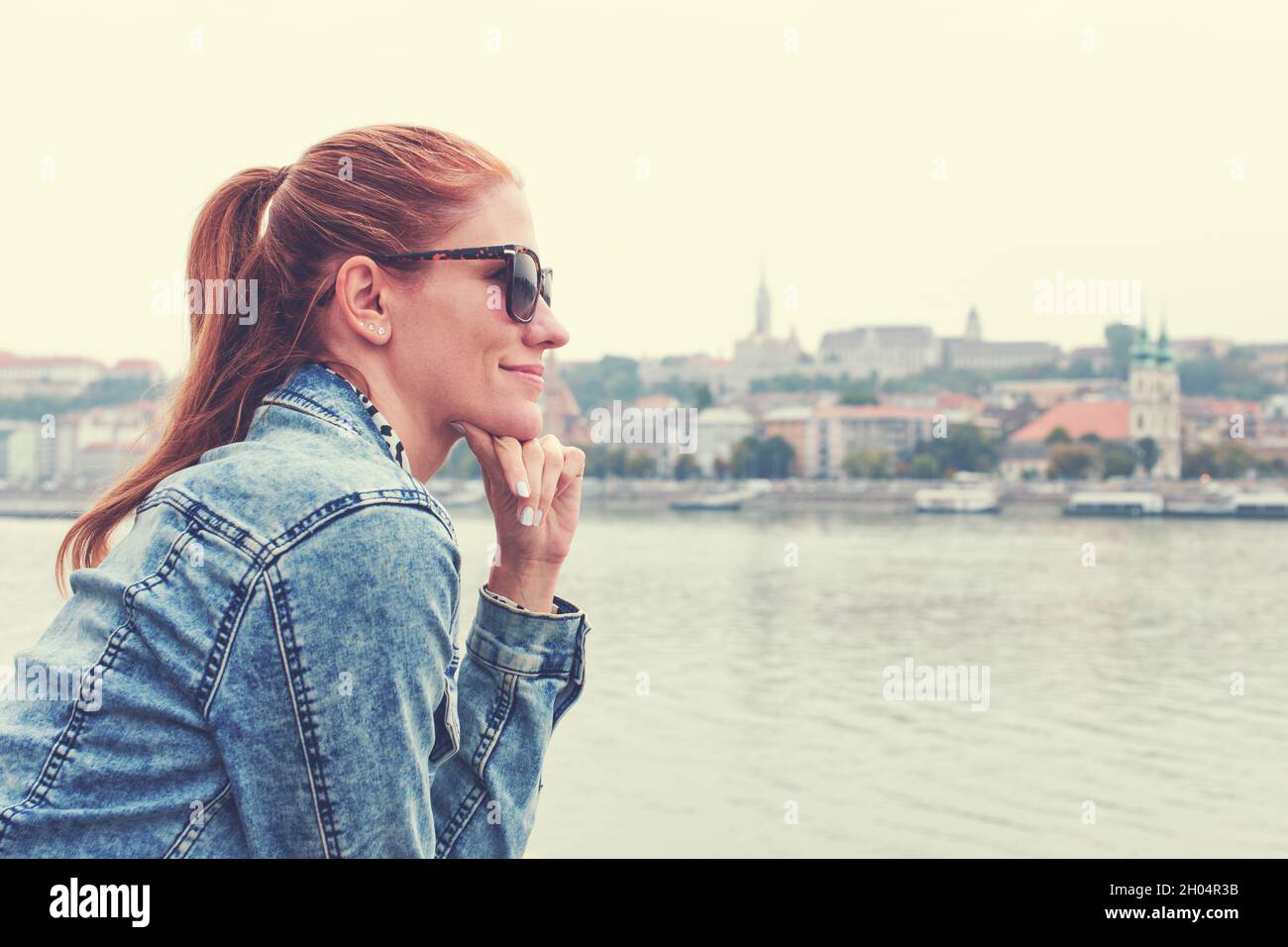 Young redhead Caucasian woman in denim jacket and sunglasses profile view at city panorama Stock Photo