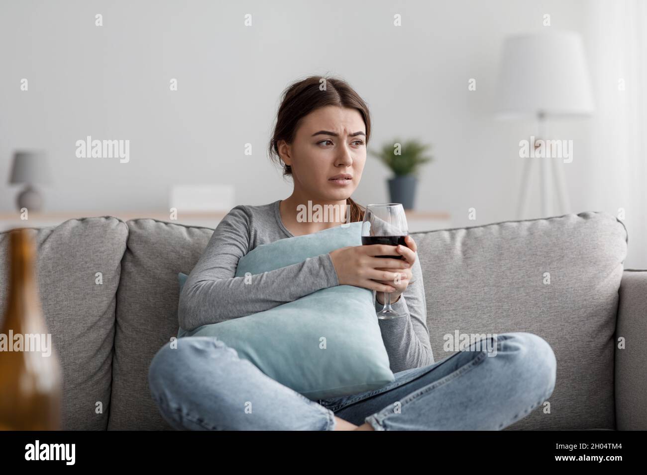 Upset apathetic european millennial woman sits on sofa with glass and suffering from depression and stress at home interior. Female alcoholic drink wi Stock Photo
