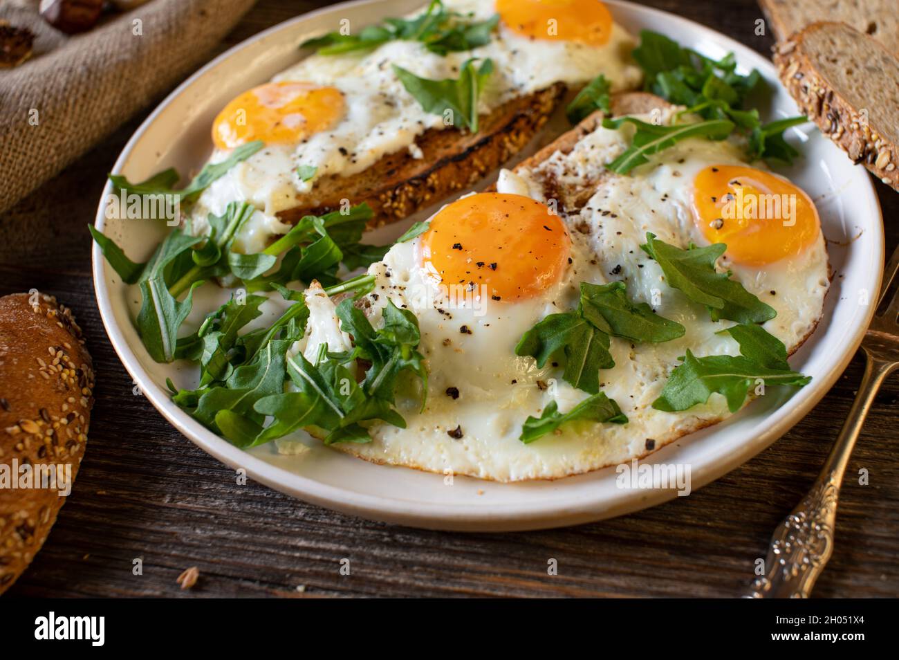 Rustic breakfast with fried eggs, sunny side up, with roasted bread and arugula. Served as a open faced sandwich on a plate on wooden table. Stock Photo