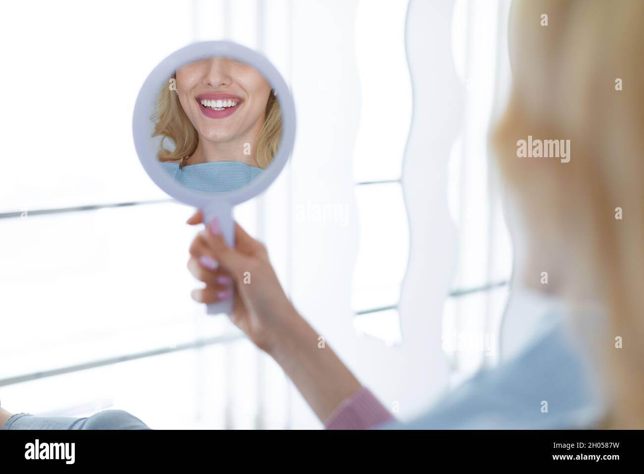 Female patient looking at her beautiful healthy white smile in dental mirror reflection, unrecognizable happy young lady checking results of treatment Stock Photo