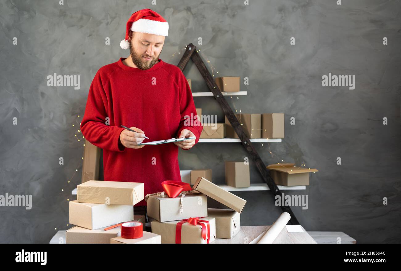 Caucasian bearded man wearing santa claus hat and red sweater business owner working at home. christmas gift shopping concept.marks boxes for delivery Stock Photo