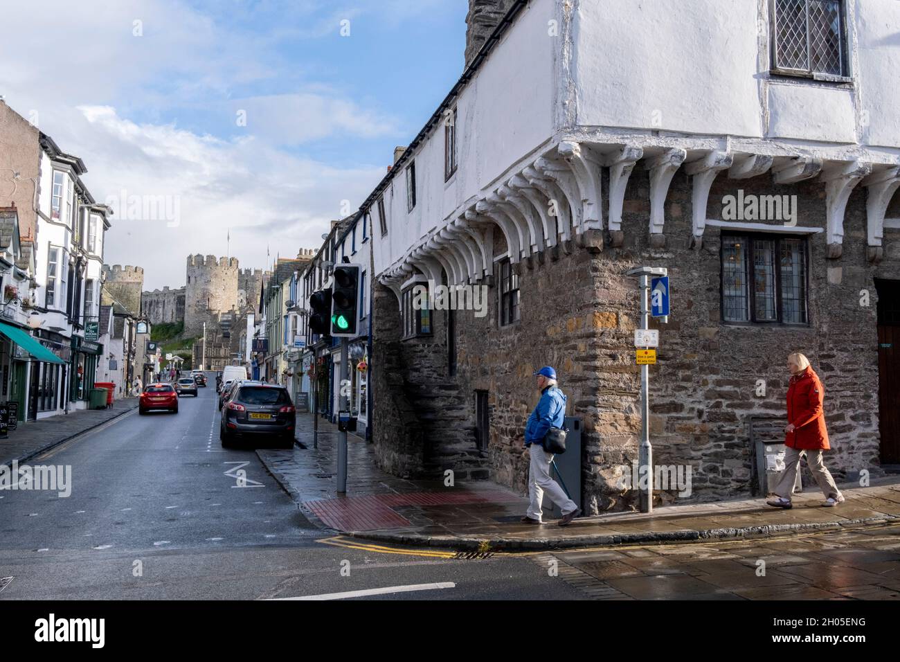 With the medieval castle dominating the town, a couple walk past the 14th century Merchant's House on Conwy high street, on 4th October 2021, in Conwy, Gwynedd, Wales. The walls were constructed between 1283 and 1287 after the foundation of Conwy by Edward I, and were designed to form an integrated system of defence alongside Conwy Castle. The walls are 1.3 km (0.81 mi) long and include 21 towers and three gatehouses. Conwy is a walled market town and community on the north coast of Wales. Stock Photo