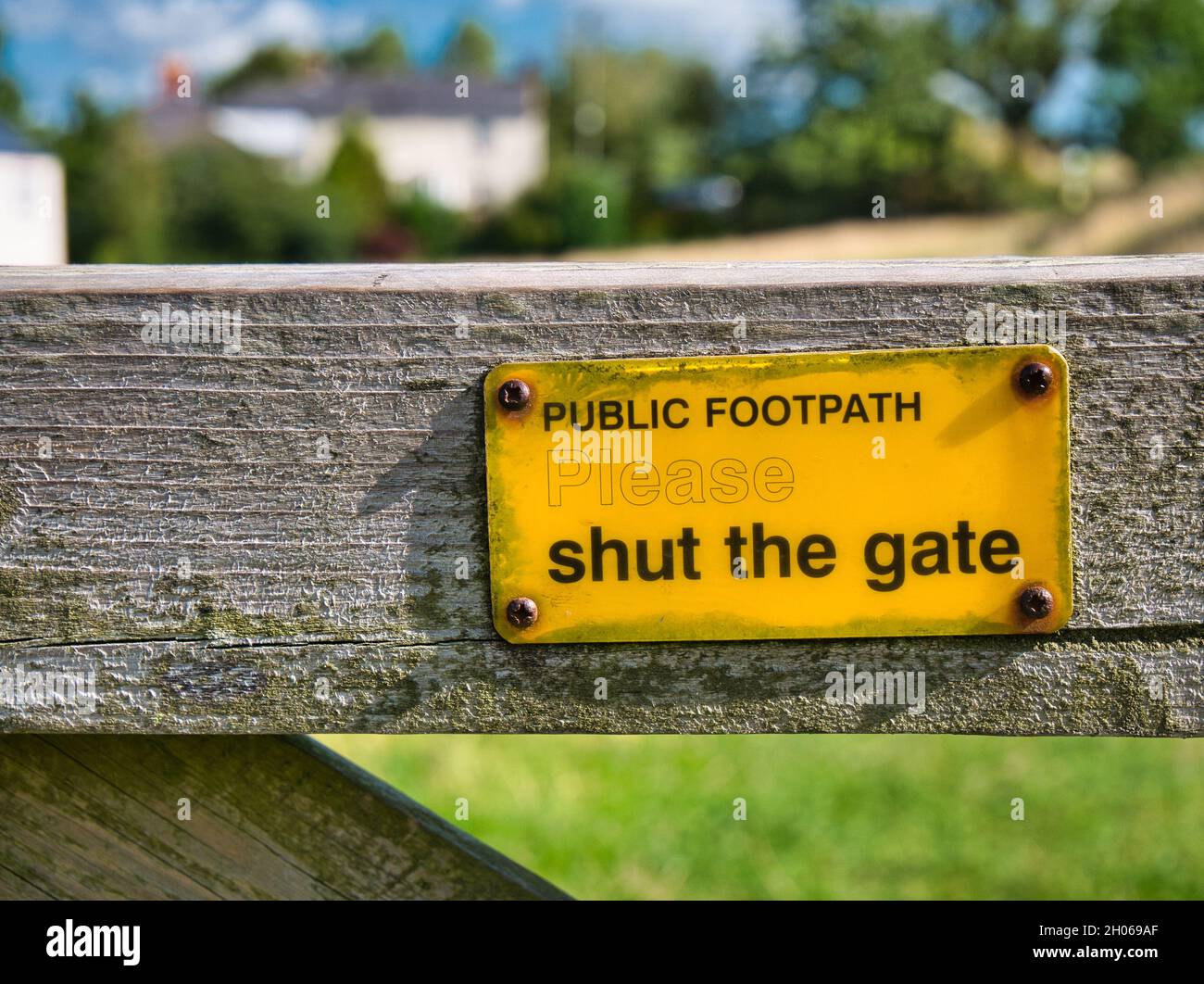 A rectangular, yellow, metal sign screwed to a wooden gate across a public footpath asks walkers to shut the gate after they pass. Stock Photo