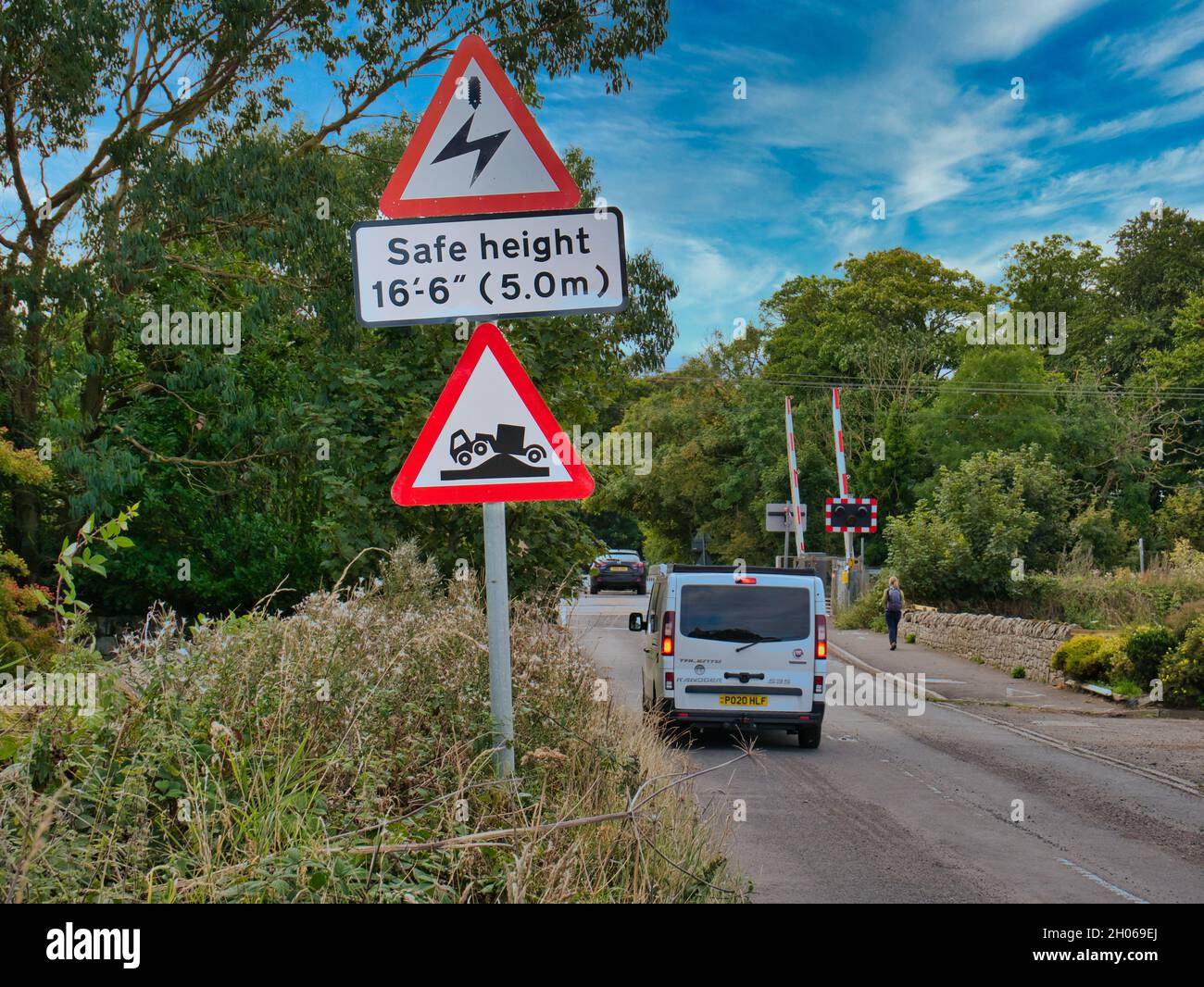 Vehicles pass signs warning of overhead electric cables and a risk of grounding as they approach a level crossing in Beal, Northumberland, UK. Stock Photo