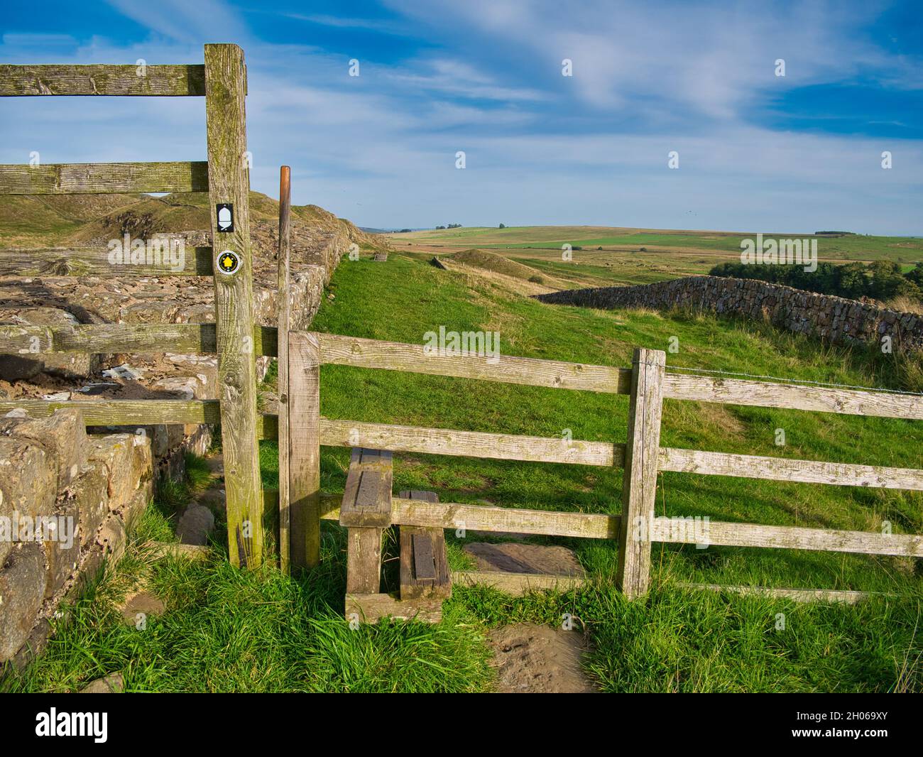 A wooden step stile with signs crosses a fence between two fields on the Hadrian's Wall Path in Northumberland, England, UK. Taken on a sunny day with Stock Photo